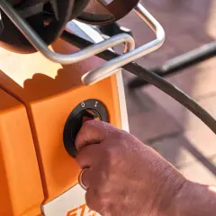 Hand dreht Schalter am orangefarbenen Hochdruckreiniger, bereit, ihn an den Wasserhahn anzuschließen.