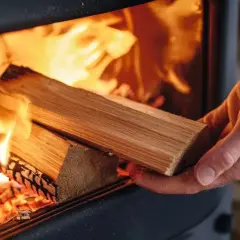Hand legt Holzscheite in einen Ofen, um feuchtes Holz zu verbrennen. Flammen lodern hell.
