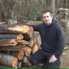 Mann steht neben einem Stapel Holzscheite. Er erklärt, dass Kiefer gutes Brennholz ist.