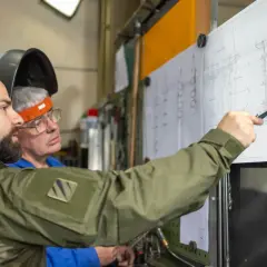 Zwei Männer besprechen einen technischen Plan. Einer zeigt mit einem Stift auf Details, die an einen **Ratschenzug** erinnern.