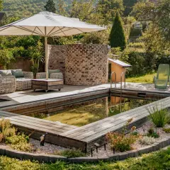 Ein schöner Garten mit einem teil-eingegrabenen Holzpool, Liegestühlen und einer gemütlichen Lounge-Ecke unter einem Sonnenschirm.