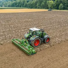Grüner Traktor mit einem breiten kreiselgrubber bearbeitet ein feuchtes Feld. Im Hintergrund ein weiteres Feld und Wald.