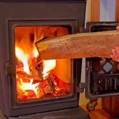 Hand legt Holzscheit in brennenden Ofen. Achtung: welches Holz darf man nicht verbrennen?