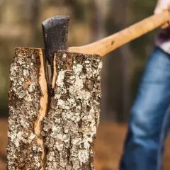 Axt steckt tief in einem gespaltenen Baumstamm. Eine Person in Jeans und kariertem Hemd bereitet sich auf den nächsten Schlag vor, um den Baumstamm zu spalten.