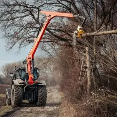 Ein Ladekran-Traktor mit orangefarbenem Ausleger schneidet einen Baumstamm in einem Wald.