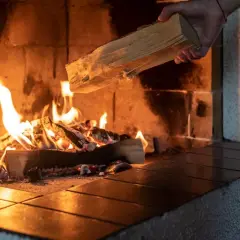Eine Hand legt Holz nach, das im Kaminfeuer brennt. Das Holz brennt nicht, es spendet Wärme.