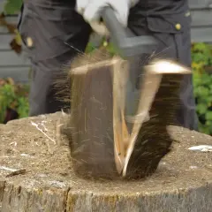Ein Mann spaltet mit einem Keil einen Baumstamm. Holzsplitter fliegen durch die Luft.
