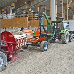 Gabelstapler lädt Holz auf eine hydraulische Kreissäge. Ein Traktor mit Anhänger wartet daneben.