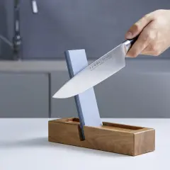 Ein Messer wird auf einem Schleifstein geschärft, der in einem Holzständer steckt. Dieser Schleifstein ist ein wichtiger Helfer, um die Schneide scharf zu halten, fast wie ein **dichtungsschaber** für die Klinge.