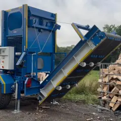 Ein Förderband für Holz transportiert gespaltene Holzscheite in einen Gitterbox-Container. Ein Traktor steht im Hintergrund.
