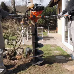 Zwei Personen bedienen einen leistungsstarken **Erdhandbohrer** im Garten. Das Gerät mit seinem orangefarbenen Motor und der langen Bohrschnecke ist bereit, Löcher für Pfosten oder Pflanzen zu graben.