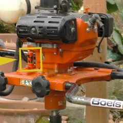 Ein orangefarbener Erdbohrer im Garten, bereit für den Einsatz. Ein gelber Pfeil hebt einen roten Hebel hervor.