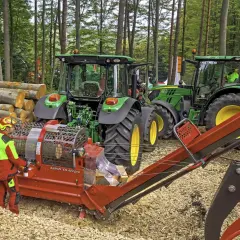 Waldarbeiter bedient einen Hacker, der Holz für die Weiterverarbeitung zerkleinert. Traktoren stehen bereit.