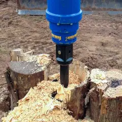 Ein blauer Bohrer entfernt einen Baumstumpf, indem er ihn zerkleinert. Holzspäne fliegen überall herum.