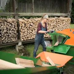 Frau arbeitet mit einer grünen und orangen Wippsäge, um Holzscheite zu schneiden. Im Hintergrund sind Holzstapel zu sehen.