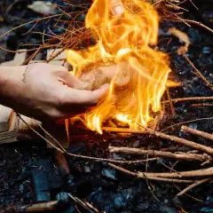 Hand zündet mit einem Feuerzeug ein Bündel Zunder an, um Wurzelholz zu verbrennen. Flammen lodern hell.