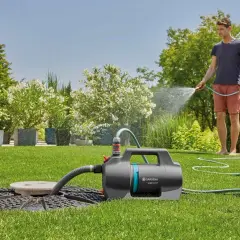 Mann bewässert den Garten mit einer Tauchpumpe, die aus einem Schacht saugt. Üppige Pflanzen und ein gepflegter Rasen.