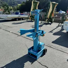 Blauer, gebrauchter Holzspalter mit Rädern steht auf einem Parkplatz. Im Hintergrund landwirtschaftliche Maschinen und Anhänger.