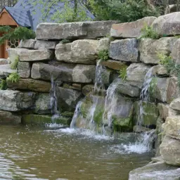 Ein kleiner Wasserfall, der wie von selbst gebaut wirkt, plätschert in einen Teich. Dahinter ein kleines Holzhaus.