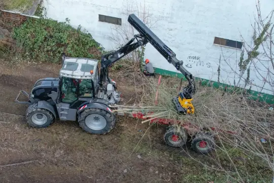 Ein schwarzer Traktor mit Kran lädt Äste auf einen Anhänger. Ein Mann in orangefarbener Kleidung arbeitet am Kran.