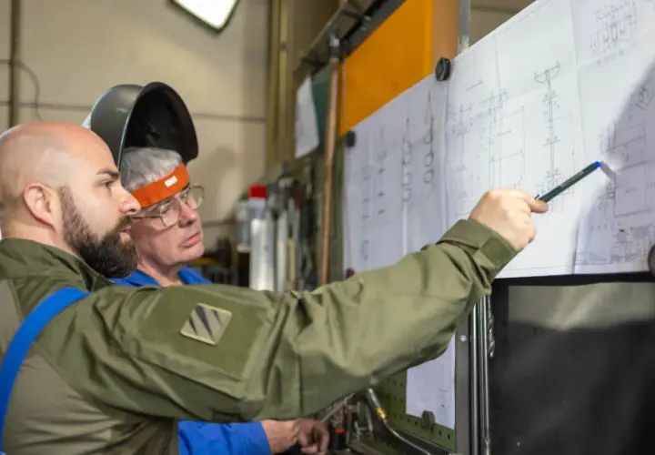 Zwei Männer besprechen einen technischen Plan. Einer zeigt mit einem Stift auf Details, die an einen **Ratschenzug** erinnern.