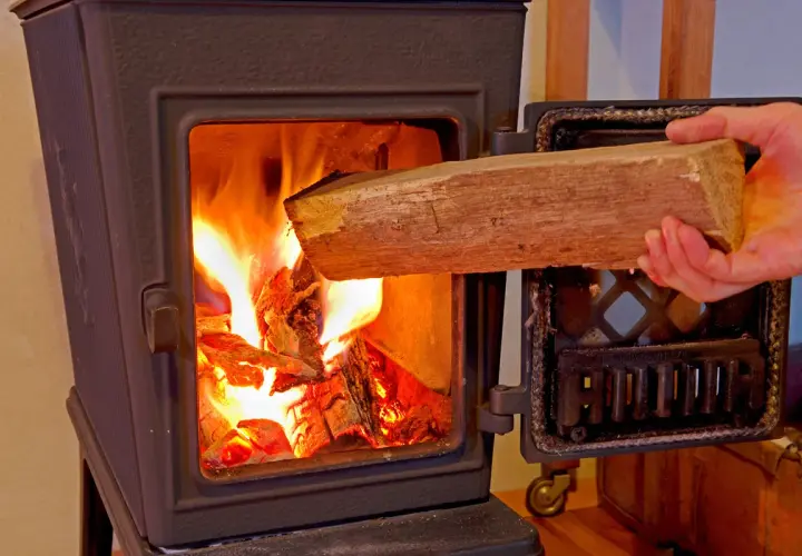 Hand legt Holzscheit in brennenden Ofen. Achtung: welches Holz darf man nicht verbrennen?