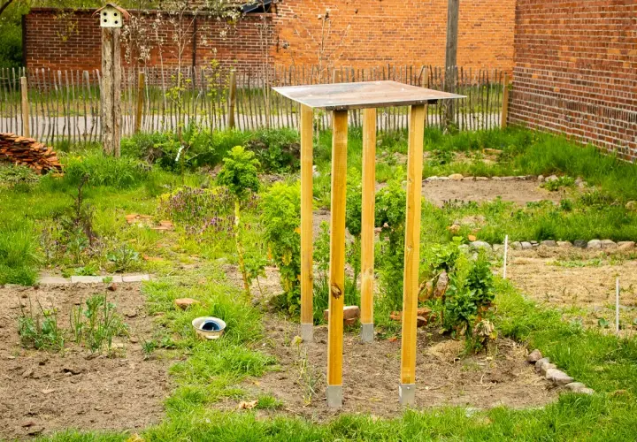 Ein provisorisches tomatendach selber bauen: Holzpfosten mit Metallfüßen tragen eine einfache Dachkonstruktion im Garten.