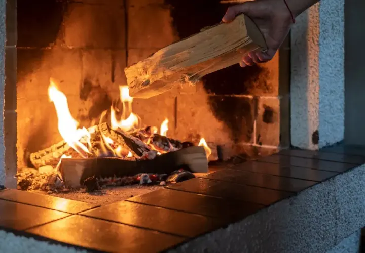 Eine Hand legt Holz nach, das im Kaminfeuer brennt. Das Holz brennt nicht, es spendet Wärme.
