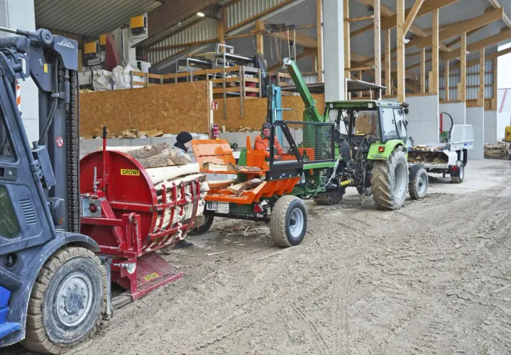 Gabelstapler lädt Holz auf eine hydraulische Kreissäge. Ein Traktor mit Anhänger wartet daneben.