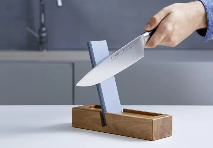 Ein Messer wird auf einem Schleifstein geschärft, der in einem Holzständer steckt. Dieser Schleifstein ist ein wichtiger Helfer, um die Schneide scharf zu halten, fast wie ein **dichtungsschaber** für die Klinge.