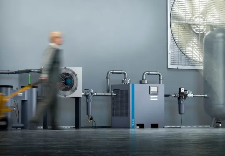 Industrielle Anlage mit Kompressor, Trockner und Luftbehälter. Ein Mitarbeiter mit Hubwagen im Vordergrund. Regelmäßiges kompressor wasser ablassen ist wichtig für die Effizienz.