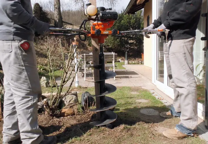 Zwei Personen bedienen einen leistungsstarken **Erdhandbohrer** im Garten. Das Gerät mit seinem orangefarbenen Motor und der langen Bohrschnecke ist bereit, Löcher für Pfosten oder Pflanzen zu graben.