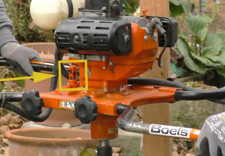 Ein orangefarbener Erdbohrer im Garten, bereit für den Einsatz. Ein gelber Pfeil hebt einen roten Hebel hervor.