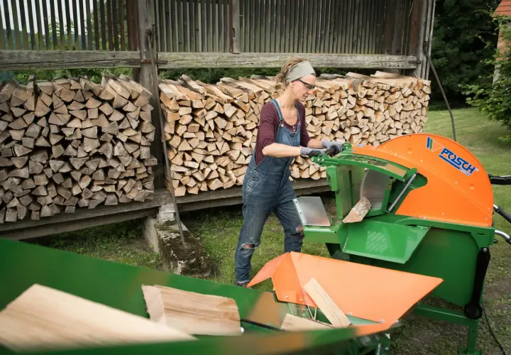 Frau arbeitet mit einer grünen und orangen Wippsäge, um Holzscheite zu schneiden. Im Hintergrund sind Holzstapel zu sehen.