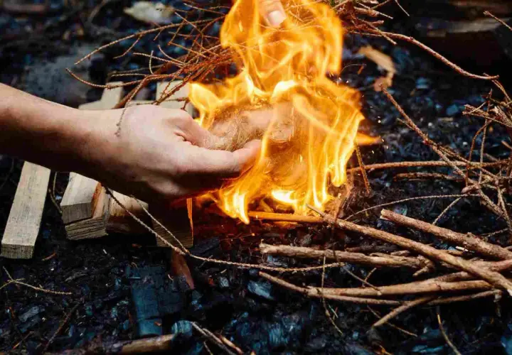 Hand zündet mit einem Feuerzeug ein Bündel Zunder an, um Wurzelholz zu verbrennen. Flammen lodern hell.