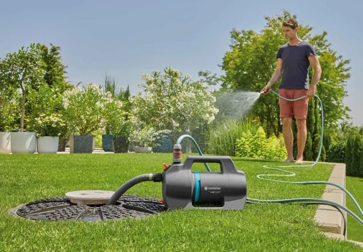 Mann bewässert den Garten mit einer Tauchpumpe, die aus einem Schacht saugt. Üppige Pflanzen und ein gepflegter Rasen.