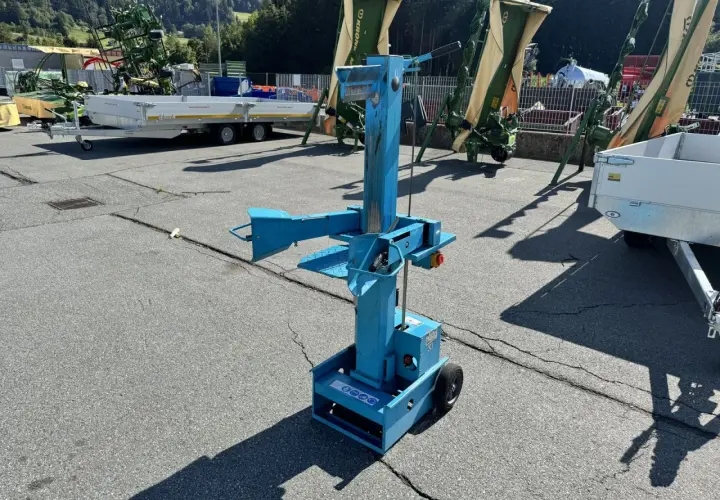 Blauer, gebrauchter Holzspalter mit Rädern steht auf einem Parkplatz. Im Hintergrund landwirtschaftliche Maschinen und Anhänger.