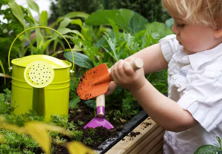 Kleines Kind mit Schaufel im Garten, lernt, wie man ein kinder hochbeet selber baut. Grüne Gießkanne daneben.