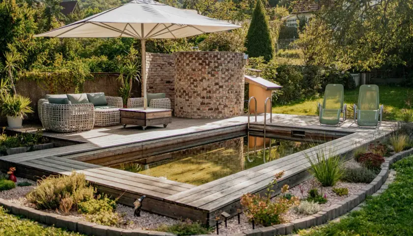 Ein schöner Garten mit einem teil-eingegrabenen Holzpool, Liegestühlen und einer gemütlichen Lounge-Ecke unter einem Sonnenschirm.