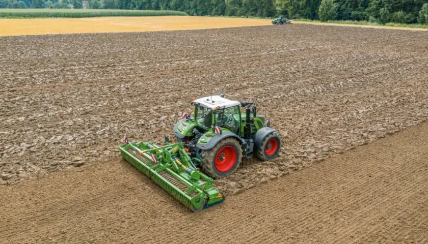 Grüner Traktor mit einem breiten kreiselgrubber bearbeitet ein feuchtes Feld. Im Hintergrund ein weiteres Feld und Wald.