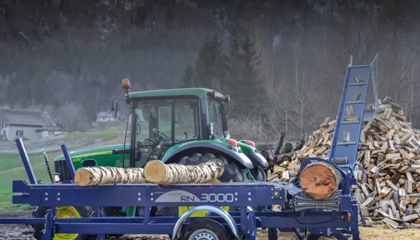 Ein Taifun Holzspalter mit Traktor im Hintergrund, bereit, einen großen Holzstapel zu verarbeiten.