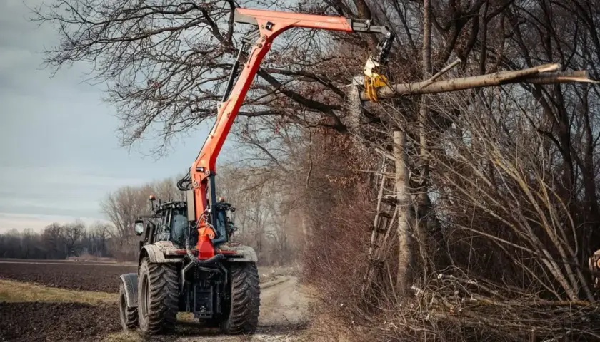 Ein Ladekran-Traktor mit orangefarbenem Ausleger schneidet einen Baumstamm in einem Wald.
