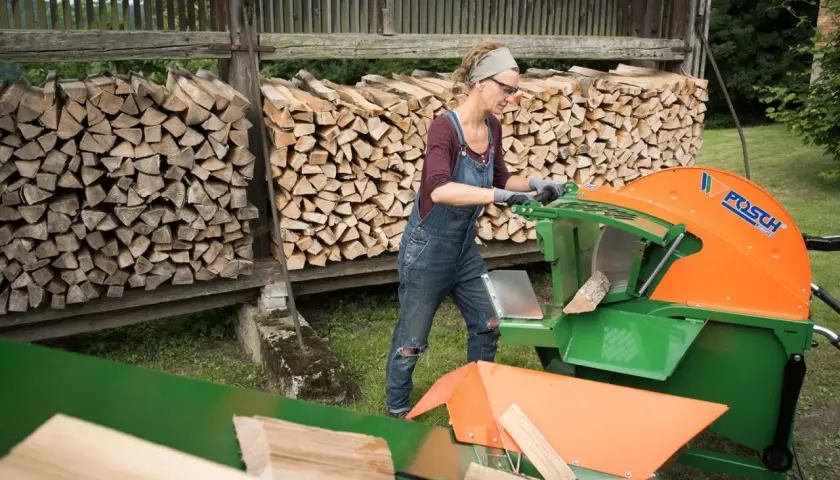 Frau bedient eine Schiebetisch Kreissäge für Brennholz, um Holzscheite zu schneiden. Im Hintergrund sind Holzstapel zu sehen.