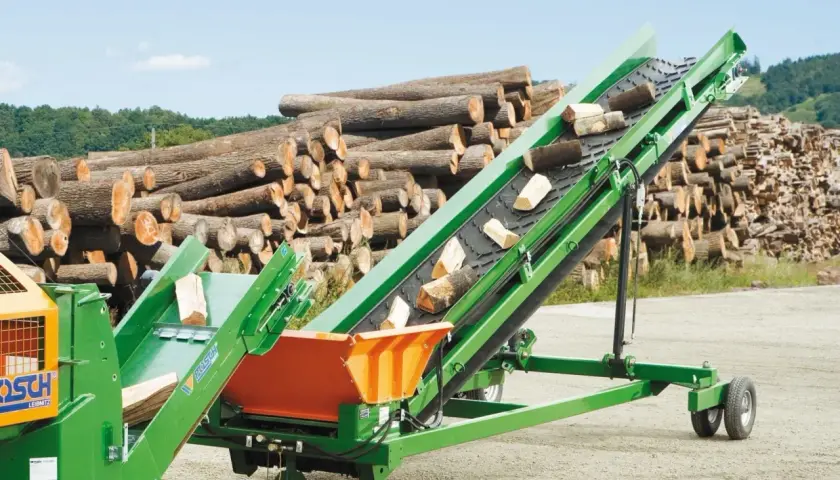 Grünes Förderband transportiert Holzscheite. Im Hintergrund liegt ein großer Stapel Baumstämme.