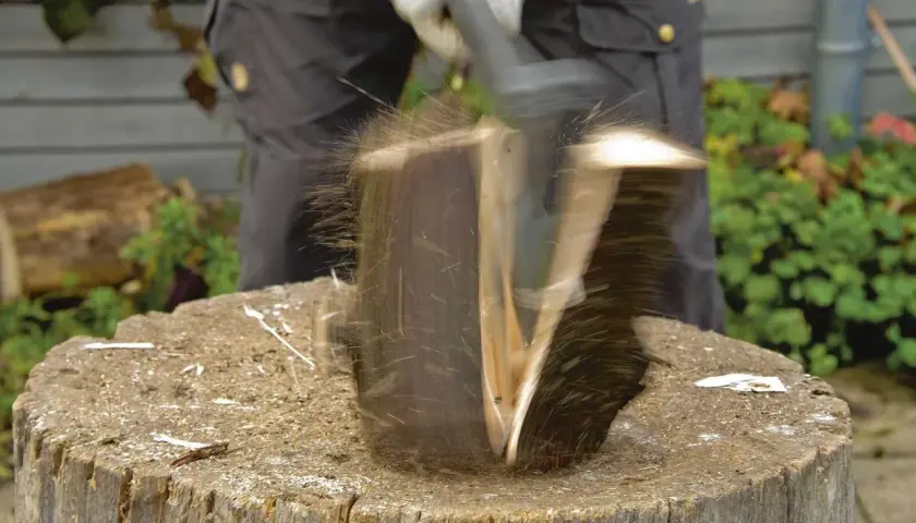 Ein Mann spaltet mit einem Keil einen Baumstamm. Holzsplitter fliegen durch die Luft.