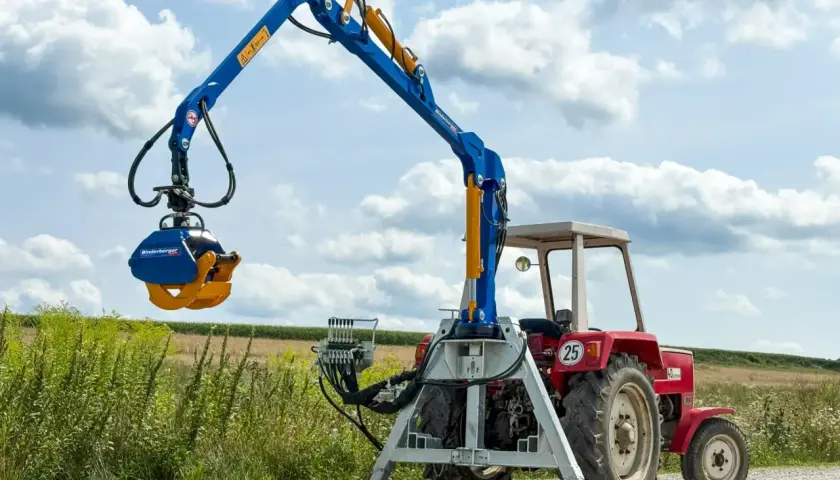 Ein blauer Forstkran mit Dreipunkt-Aufnahme und Greifer steht neben einem roten Traktor im Feld.