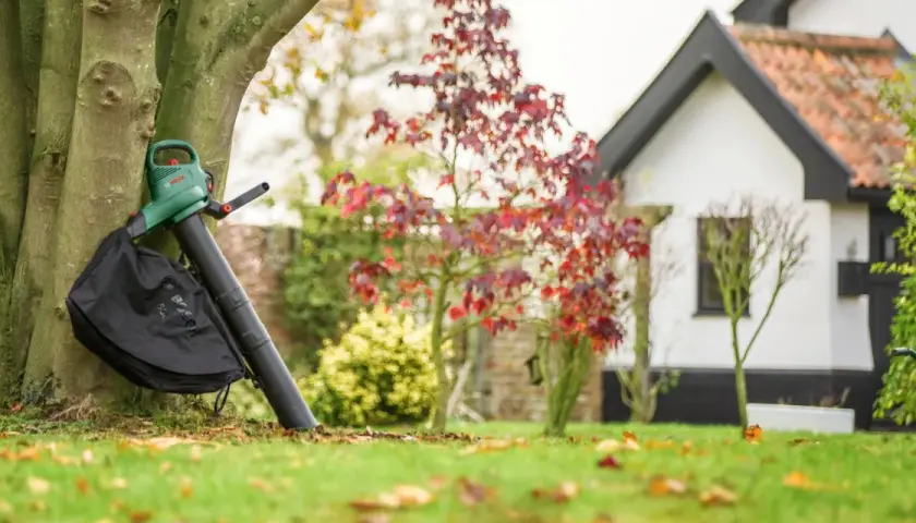 Laubbläser mit Fangbeutel lehnt am Baum, bereit, das Herbstlaub zu zerkleinern. Im Hintergrund ein Haus und Garten.