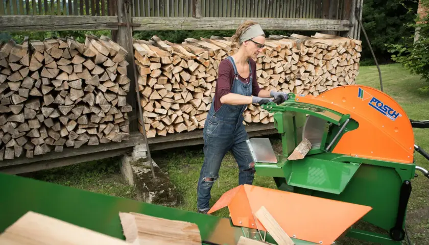 Frau arbeitet mit einer grünen und orangen Wippsäge, um Holzscheite zu schneiden. Im Hintergrund sind Holzstapel zu sehen.