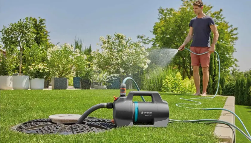 Mann bewässert den Garten mit einer Tauchpumpe, die aus einem Schacht saugt. Üppige Pflanzen und ein gepflegter Rasen.