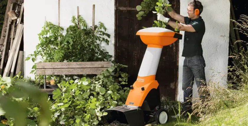 Ein Mann mit Schutzbrille und Gehörschutz bedient einen orange-weißen Holzhacker. Die Zapfwelle des Geräts ist bereit für den Einsatz im Garten.